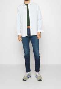 Bílé tričko s dlouhým rukávem přes zelené tričko, modré džíny a šedé tenisky s modrými detaily. Oblečení doplňuje hnědý pásek.