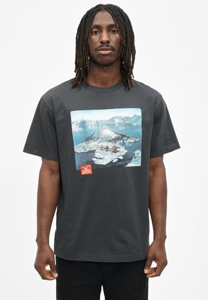 Man with shoulder-length dreadlocks wears dark gray T-shirt showing snowy mountain landscape with small people and red logo.