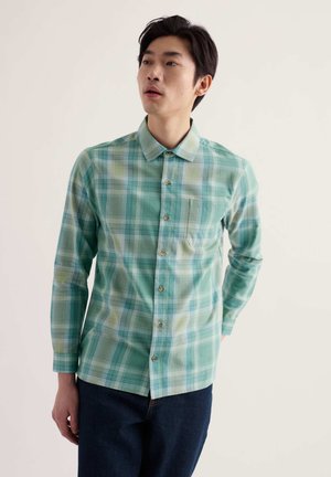 Homme portant une chemise à carreaux verte et bleue à manches longues et un pantalon sombre, debout devant un fond clair uni.