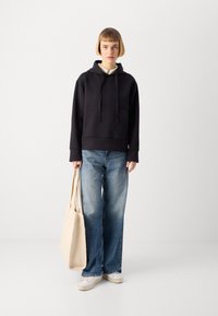 Felpa nera in cotone con tasca frontale, abbinata a jeans blu sbiaditi e larghi e scarpe da ginnastica bianche. Tiene una borsa in tessuto beige.