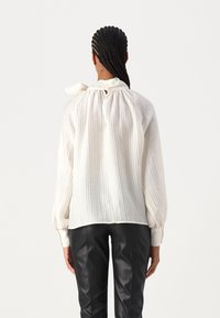 Personne avec des cheveux tressés portant un blouse blanc texturé avec un nœud au cou et un pantalon en cuir noir, vue de dos sur un fond uni.
