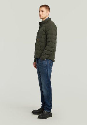 Veste matelassée verte foncée avec une fermeture éclair, des coutures verticales, un col monté et un accent de logo. Associée à un jean bleu et des bottes noires.