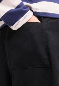 Χέρι στην τσέπη μαύρου παντελονιού φορεμένου με πουκάμισο μακρυμάνικο με οριζόντιες μπλε και λευκές ρίγες.