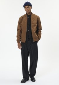 Chaqueta bomber marrón, camisa negra abotonada, pantalones negros y zapatos negros. El modelo lleva un gorro de punto azul y posa sobre un fondo blanco.