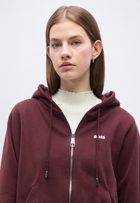 Jeune femme aux cheveux bruns raides portant des boucles d'oreilles argentées en forme d'anneau, un haut blanc côtelé et un sweat à capuche bordeaux zippé avec le logo "BOSS".