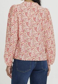Blouse à fleurs avec un fond beige clair, ornée de motifs rouges et jaunes. Manches bouffantes, encolure froncée et poignets élastiques.