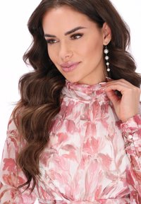 Bloes met bloemendruk in sheer, licht materiaal met tinten roze. Hoge kraag en knoopdetails op de mouwen. Lang, golvend haar en pareloorbellen.