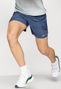 Marineblaue Sportshorts mit einem leichten, glatten Material; ausgestattet mit einem seitlichen Schlitz und einem reflektierenden Nike-Logo. Kombiniert mit weißen Sneakern.