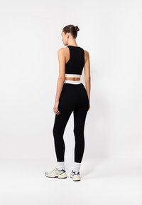 Kobieta stojąca tyłem, ubrana w czarny bezrękawnik, czarne legginsy z białym pasem w talii, białe skarpetki i białe trampki.