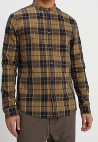 Brown and navy plaid shirt with a band collar, long sleeves, and blue buttons. Made of woven fabric with a smooth texture.