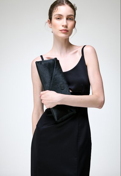 Femme en robe noire sans manches tenant une pochette noire en fourrure sur un fond gris clair neutre.