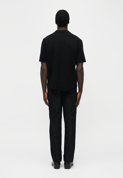 Άνδρας που στέκεται γυρισμένος προς τα πίσω, φοράει μαύρο πουκάμισο με κοντά μανίκια, μαύρο παντελόνι και μαύρα παπούτσια, σε απλό φωτεινό φόντο.