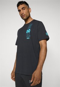 Sort bomulds T-shirt med korte ærmer. Har turkise tekster "JUST DO IT" og "INTER" lodret ned langs venstre side, med et logo på ærmet.