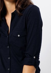 Navyblauw button-up shirt, lange mouwen opgerold, twee borstzakken, zilveren knoopaccenten, zachte stoftextuur, subtiel gemêleerd patroon.
