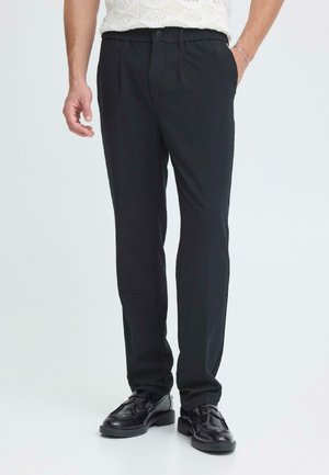 Schwarze maßgeschneiderte Hose mit glatter Textur, ausgestattet mit einem Knopfverschluss und seitlichen Taschen, kombiniert mit schwarzen Lederschuhen.