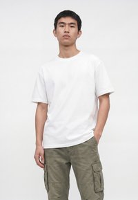 Junger Mann trägt ein schlichtes weißes kurzärmeliges T-Shirt und olivgrüne Cargohosen, steht mit neutralem Gesichtsausdruck und einer Hand in der Tasche.