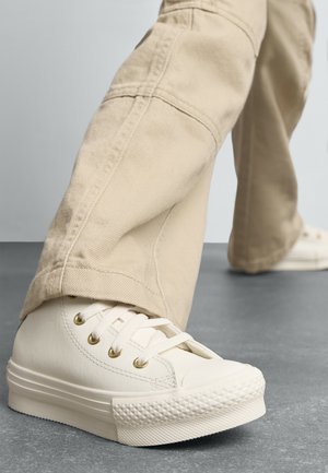 Witte leren sneakers met gouden ogen en een getextureerde rubberen zool, gecombineerd met beige broek. Focus op de details van de schoenen en de broek, minimale achtergrond.