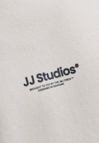 Helles beige Gewebe mit strukturierter Strickoberfläche. Bedrucktes schwarzes Logo mit der Aufschrift "JJ Studios" und kleinerem Text darunter. Einfaches, klares Design.