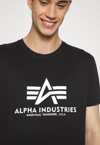 Black cotton t-shirt featuring a large white logo with an 'A' and three horizontal stripes. Round neckline and short sleeves.