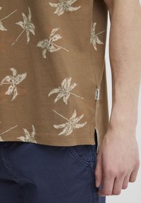Barna, rövid ujjú ing fehér levélmintákkal, oldalvágásokkal és címkével. Sötétkék nadrág felett viselve. Texturált anyag, természetes megjelenéssel.