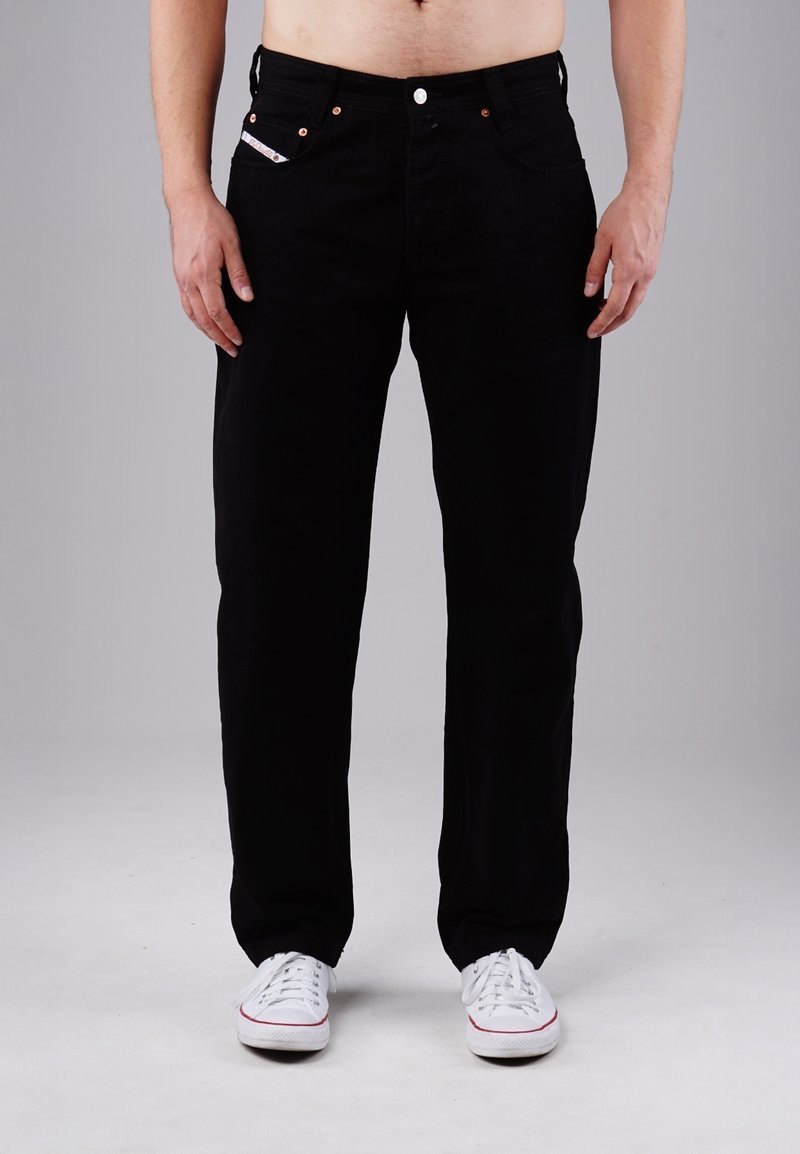 Schwarze Denim-Jeans mit geradem Schnitt, Fünf-Taschen-Design, kupferfarbenen Nieten und tonalem Nähen, kombiniert mit weißen Turnschuhen.