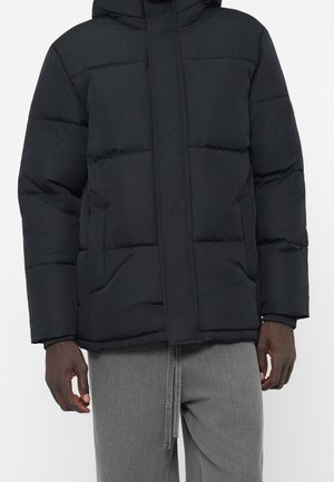 Doudoune noire matelassée avec capuche et poches avant portée par une personne en pantalon gris, visible du cou aux hanches.