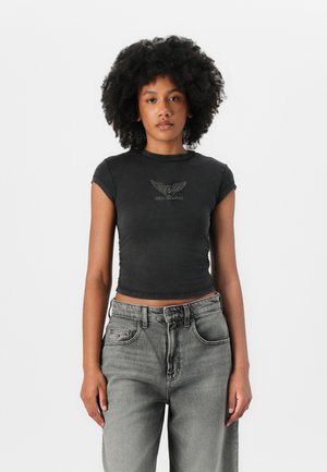 Juodas trumpas marškinėliai su trumpomis rankovėmis, ant kurių yra sparnuotas logotipas ir tekstas "GUESS ORIGINALS" sidabrinės spalvos. Sukrauti su šviesiai pilkų aukštu liemeniu džinsais.