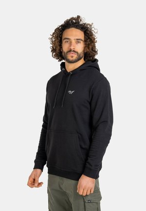 Mann mit lockigem Haar, der einen schwarzen Hoodie und olivgrüne Cargohosen trägt, steht vor einem schlichten hellen Hintergrund.