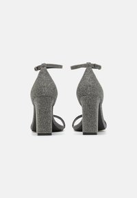 Grå teksturerte høyhælte sko med blokkhel, ankelremmer og åpen tå-design. Materialet har en subtil glitrende effekt.