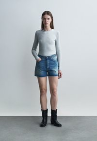 Tânără în picioare, purtând o bluză cu mâneci lungi, de culoare gri deschis, pantaloni scurți din denim albastru și cizme negre până la gleznă, în fața unui fundal simplu.