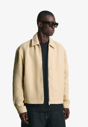 Beige textured jacket with a collared neckline, full zipper, and relaxed fit, paired with a black t-shirt and sunglasses.