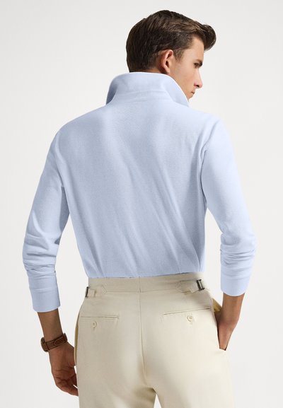 Άντρας που φοράει γαλάζιο πουκάμισο με μακριά μανίκια και ανασηκωμένο γιακά, με μπεζ παντελόνι, στέκεται με το ένα χέρι στην τσέπη, γυρισμένος προς τα πίσω.