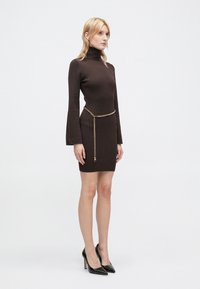 Brun ribbet strikkjole med høj hals og klokkeærmer, accentueret af et guld kædebælte, parret med sorte højhælede sko.