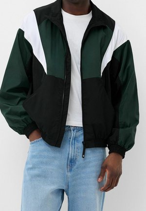 Mand iført sort, mørkegrøn og hvid color-block lynlåsjakke over en hvid T-shirt og lyseblå jeans, hænderne i lommerne.