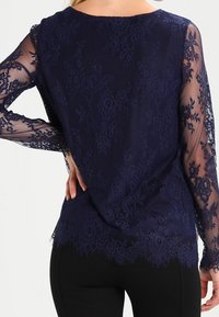 Blusa in pizzo blu navy con maniche lunghe trasparenti, orlo scallop, motivo floreale e scollatura rotonda. Dettagli testurizzati a strati sono visibili.