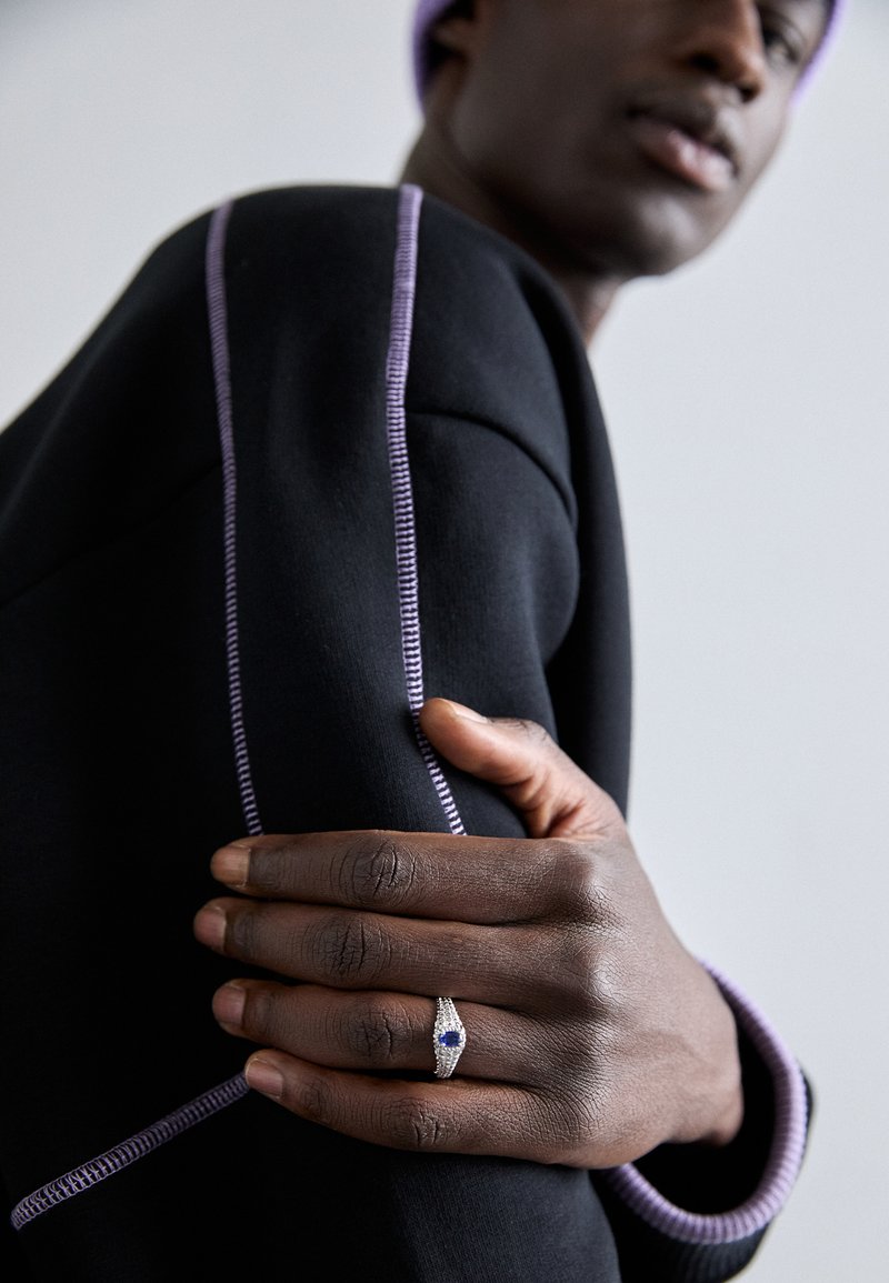 Črna majica s kapuco in vijoličnim šivom; roka, okrašena s srebrnim prstanom z osrednjim modrim kamnom, počiva na rokavu.