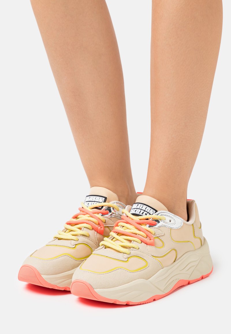 Beige sneakers with textured fabric, neon yellow accents, pink soles, and colorful laces; rounded shape and padded collar.