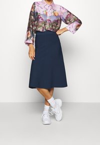Hemd mit Blumen- und Obstdruck in Lila, kombiniert mit einem knielangen marineblauen Rock. Weiße klobige Sneakers und Socken runden den Look ab.