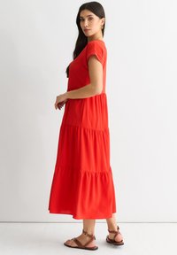 Red, tiered maxi dress with short sleeves, flowing fabric, and a relaxed fit. Paired with brown strappy sandals. Simple design, no patterns.