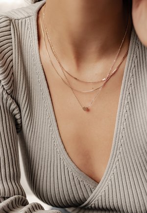Three layered gold necklaces featuring varying chain styles, including a heart-shaped pendant, worn over a ribbed grey top.