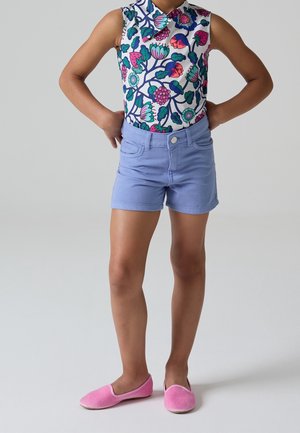 Barn iført en ærmeløs blomstret top, lyseblå shorts og pink slip-on sko, stående med hænderne på hofterne på ensfarvet baggrund.