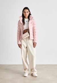 Ανοιχτό ροζ φουσκωτό μπουφάν με οριζόντιες ραφές, κουκούλα, φορεμένο πάνω από ένα λευκό cropped top και καφέ φαρδιά παντελόνια. Λευκά αθλητικά παπού�τσια και μια μικρή τσάντα.