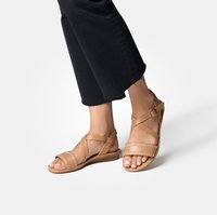 Sandales en cuir tan avec des sangles croisées et des coutures décoratives, semelle plate et boucle de cheville ajustable. Portées avec un pantalon noir raccourci.