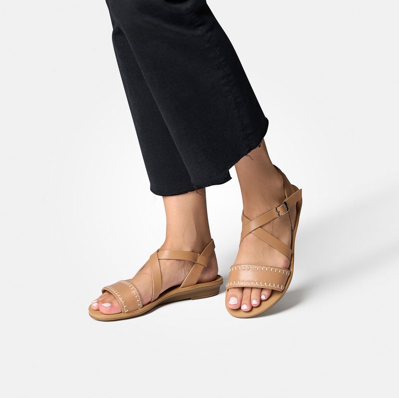 Sandales en cuir tan avec des sangles croisées et des coutures décoratives, semelle plate et boucle de cheville ajustable. Portées avec un pantalon noir raccourci.