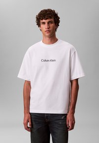 Hvit t-skjorte i bomull med korte ermer med "Calvin Klein"-logo i svart. Skjorten har en avslappet passform og rund hals. Kombinert med mørke jeans.