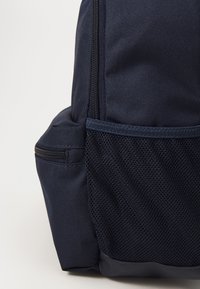Sac à dos bleu marine en tissu texturé avec une poche latérale en maille. Il comporte un compartiment zippé et un fond structuré pour la stabilité.