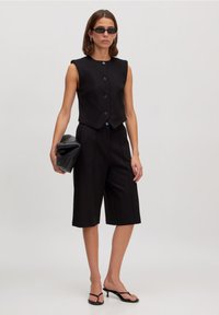 Gilet sans manches noir à cinq boutons, associé à des culottes larges noires. Les accessoires comprennent une pochette noire et des sandales minimalistes noires.
