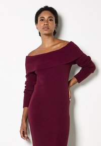 Donna con capelli ricci scuri, che indossa un vestito a maglia bordeaux attillato con maniche lunghe e scollatura a spalle scoperte, in piedi con una mano sui fianchi davanti a uno sfondo bianco.