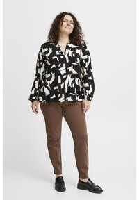 Blusa nera con motivi astratti bianchi e beige, abbinata a pantaloni marroni aderenti e mocassini neri. Il tessuto sembra liscio.