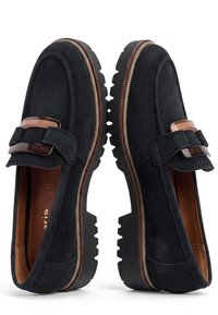 Paire de mocassins en daim noir avec semelles épaisses en caoutchouc et détail décoratif avec boucle marron sur le dessus, sur fond blanc.
